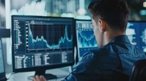 A person sits at a desk, focusing on multiple computer monitors displaying financial graphs and data charts, indicating stock market analysis. The room is dimly lit, giving an atmosphere of concentration and focus.