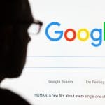 Silhouette of a person in glasses facing a computer screen displaying the Google homepage. The screen features a search bar with options "Google Search" and "I'm Feeling Lucky". Text at the bottom mentions a film titled "HUMAN".