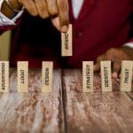 A person in a red suit arranges wooden blocks with words like "Marketing," "Logo," and "Brand" on a wooden table. The focus is on the "Brand" block, which is being placed upright among the others.