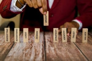 A person in a red suit arranges wooden blocks with words like "Marketing," "Logo," and "Brand" on a wooden table. The focus is on the "Brand" block, which is being placed upright among the others.